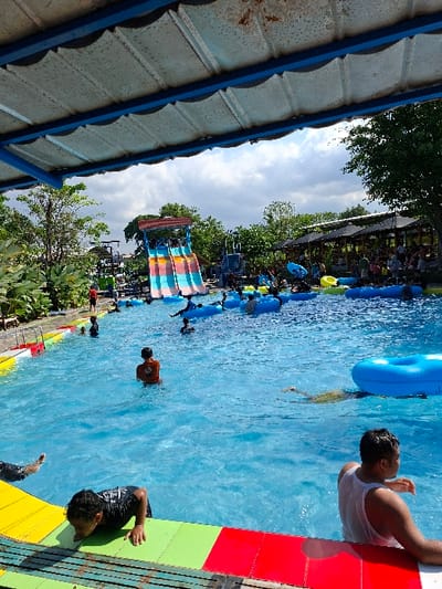 The image shows a swimming pool in Kecamatan Tulung, Indonesia, located at latitude: -7.592, longitude: 110.65. There is a blue and pink waterslide in the background, and people are swimming and playing in the pool. One man in a white tank top sits on the edge of the pool. There is a blue floating tube in the middle of the pool. The image is taken from under a roof. The pool is surrounded by green vegetation. The sky is cloudy and bright.