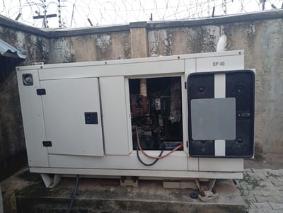 A white generator, model SP 40, is situated on a concrete base in an outdoor setting. The generator has its main access door on the left open, revealing internal machinery. The right side of the generator features a closed black panel with a white vertical stripe. The generator is positioned against a textured, weathered concrete wall topped with a security fence consisting of barbed wire and posts. To the left of the generator, a corrugated metal roof is visible. The ground surface around the generator is concrete, with some overgrowth of vegetation. The scene appears to be in Zanzi, Nigeria, given the presence of barbed wire fencing commonly used for security in such locations. There are no people present in the image.