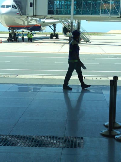 An airport scene in Bắc Cam Ranh, Vietnam, shows a commercial airplane connected to a jet bridge. In the foreground, a male airport worker wearing a dark blue uniform with green accents and a blue cap walks across a tiled floor. He is carrying a white document in his right hand. The worker's shadow is cast on the floor. Behind him, an airplane is visible, with its jet bridge extended to the aircraft. Several other workers in high-visibility vests are standing near the airplane's wheels. The environment appears to be an airport tarmac under a bright, clear sky.