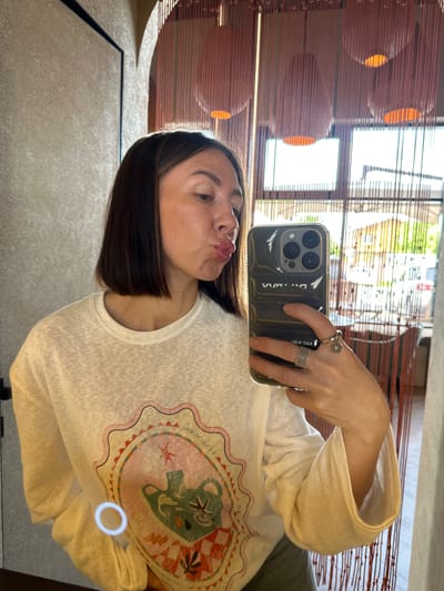 A young adult female is captured in a mirror selfie indoors, appearing to be within a commercial establishment such as a restaurant or cafe. She has medium-length, dark brown hair styled in a bob cut and is pouting her lips in a "duck face" expression, looking slightly downward and to her right. Her skin appears fair, and her lips are glossy. She is wearing a loose-fitting, off-white, long-sleeved t-shirt made of a textured (slub knit) fabric. The front of her shirt features a large graphic design within a scalloped, colorful frame. The central motif includes a stylized green teapot or pitcher, possibly with a spout resembling a bird's head, surrounded by what appears to be leaves, stars, and a checkered pattern in shades of red, white, and brown. Partial text is visible on the graphic, including "il cielo stellato" (Italian for "the starry sky") and "DI CIOCO," along with other illegible script. A small, bright circular reflection is visible on the lower left portion of her shirt. A sliver of light green or khaki fabric, possibly trousers or a skirt, is visible at the bottom edge of the frame. The woman holds a dark, possibly black or dark grey, padded or quilted smartphone case with her left hand, which holds the phone angled towards her face. The phone itself appears to be an iPhone model, identifiable by its triple rear camera array. Reversed text on the case reads "WANG" (suggesting Alexander Wang) and other partially visible, reversed text such as "DO SANTI" or "PRO 81 TOEA." Her left hand displays two rings on her thumb: a wide, multi-banded silver-toned ring, and another on her ring finger, which includes a simpler band ring and a charm ring with a small, silver-toned flower. The background reveals a stylish interior with a textured, light beige or grey wall on the left, featuring a dark vertical trim or doorframe. Further back, two arched architectural elements are visible. These arches are partially obscured by a screen or partition made of numerous reddish-brown vertical strings, beads, or thin fabric strips, creating a semi-transparent barrier. Within this screen, several large, spherical or bell-shaped light fixtures, also reddish-brown and translucent, are suspended, emitting a warm, orange glow. Beyond the screen and arches, large windows show an outdoor scene under bright daylight. Green foliage, trees, and parts of residential or commercial buildings (one with a light-colored roof) are discernible