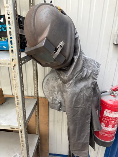 In Mol, Belgium, a welding helmet and protective suit hang on a metal rack next to a fire extinguisher. The scene appears to be an indoor storage area, possibly a workshop or industrial space, with a corrugated metal wall and industrial shelving units visible. The welding helmet is dark gray and appears to be a professional-grade model, with a visible visor and straps. The protective suit is also dark gray and made of a heavy-duty material, designed to shield the wearer from sparks and heat. The fire extinguisher is red and appears to be a standard industrial model. The overall impression is one of organized safety equipment, ready for use.
