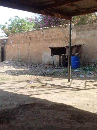 The image depicts an outdoor scene in Sabon Gari West, Nigeria, under bright daylight, viewed from beneath a corrugated metal roof structure. The foreground features a light brown dirt ground, partially obscured by prominent, elongated shadows cast by an unseen overhead structure and a vertical metal support pole located in the mid-right. Scattered white plastic bags and other refuse are visible on the sunlit dirt.

The midground is dominated by a weathered, light brown block wall with visible mortar lines, running horizontally across the frame. The wall exhibits areas of chipped plaster and discoloration. A coping of small, lighter-colored tiles or bricks caps the wall. Adjacent to the wall, right of center, stands a small, rudimentary shelter with a corrugated metal roof, supported by thin metal elements. To the immediate right of this shelter, a blue plastic barrel, bearing the white text "ST. CHARLES," rests upright against the wall. The ground area leading to the wall is heavily littered with various debris, including plastic, paper, and general refuse, interspersed with sparse, dry vegetation.

In the background, above the wall, dense green foliage is visible. A prominent bush or tree on the right displays vibrant purple or pink flowers. A sliver of clear, bright sky is visible at the top. To the far left, a dark, recessed opening, possibly a doorway or entrance, is discernible within another structure or wall beyond the main block wall. No individuals are present in the scene.