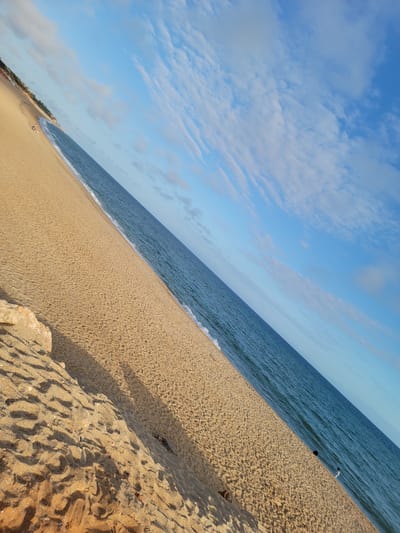 This is a photograph taken at the beach in Quarteira, Portugal. The image is taken from a low angle and is tilted, giving a sense of depth and movement. The foreground is dominated by sand, with a large rock formation on the left side of the frame. The sand is textured with footprints and small debris, and casts long shadows from an unseen light source.
The middle ground of the image is the ocean, with gentle waves lapping at the shore. The water is a deep blue, with a lighter blue horizon line where it meets the sky. A single person, wearing white clothing, is visible in the water. A black object, possibly a bag or a beach umbrella, is near the person in the water.
The background is the sky, which is a clear blue with scattered white clouds. The clouds are wispy and streaky, suggesting a breezy day. In the far distance, along the coastline, there are hints of buildings and vegetation, indicating the presence of the town of Quarteira. The overall impression is one of a peaceful and serene beach scene.