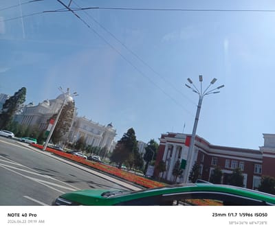 The photograph captures a street scene in Dushanbe, Tajikistan, on June 22, 2026, at 9:19 AM. The image features a bright, clear sky and is taken from a slightly elevated perspective, possibly from inside a vehicle. In the foreground, the hood of a green car is visible. The middle ground displays a street with several cars parked and in motion. Adjacent to the street is a vibrant flowerbed filled with red and orange flowers, bordered by trees.
Dominating the background are two prominent buildings. On the left, a large, ornate white building with classical architectural elements, including columns and domes, is visible. A tall lamppost with multiple lights stands in front of this building, near a flagpole displaying the flag of Tajikistan. To the right of the white building is a reddish-brown building with a similar classical design, featuring columns and a prominent white roofline. The overall environment suggests a well-maintained urban setting. The GPS coordinates provided are 38°34'26"N 68°47'28"E.