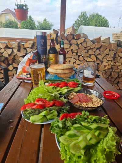 A still life image captures a rustic outdoor dining setting on a wooden table, likely in the city of Gornye Moriny, Russia, given the presence of stacked firewood and a greenhouse visible in the background. A platter of fresh vegetables, including lettuce, tomatoes, cucumbers, and dill, is arranged on the table, accompanied by a bowl of peanuts and sliced salami. Two glasses of beer, a bottle of vodka, and a bottle of some darker liquid are also present, along with a stack of bread and a bag of chips. The scene is set against a backdrop of a clear sky and a residential building, suggesting a casual gathering or meal enjoyed in a backyard or outdoor patio.