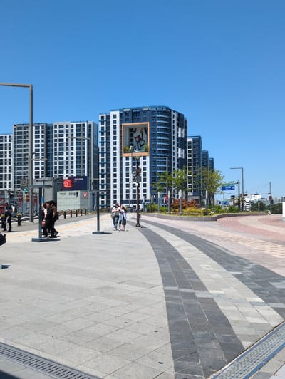 The image captures an urban landscape in Daegu, South Korea, featuring modern high-rise apartment buildings against a clear blue sky. The buildings are primarily white and gray with numerous windows. A large billboard is visible on one of the buildings.
A wide pedestrian walkway dominates the foreground, paved with light-colored concrete tiles. Along the walkway, there are benches with people sitting, standing, and interacting. The walkway transitions to a brick-paved area with alternating gray and brown brick patterns. Streetlights and trees line the sidewalk adjacent to the buildings.
In the distance, more buildings and street infrastructure are visible, including traffic lights and street signs written in Korean. The overall environment is clean and well-maintained, suggesting a modern urban area.
