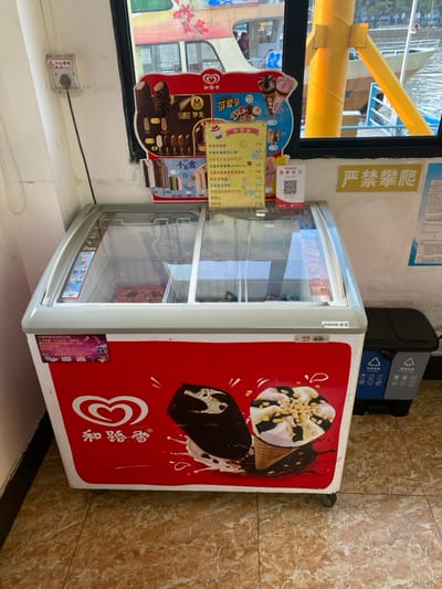 The media file, rotated clockwise by 90 degrees, captures an indoor scene, likely a small shop or kiosk in Guangzhou City, China, situated next to a body of water.

The main subject is a white ice cream display freezer with a glass top, featuring a prominent red side panel adorned with large illustrations of ice cream cones and bars, alongside the "Wall's" (和路雪) brand logo and a heart icon. The word "HIRONB" is also visible on the freezer's top edge. Attached to the freezer, on the left, is a colorful menu board displaying various ice cream options, including "可爱多" (Cornetto) and "梦龙" (Magnum), with prices listed in Chinese Yuan (e.g., ¥4, ¥6, ¥8, ¥12, ¥16, ¥18). A QR code is present on the menu board, likely for payment or information. The glass display section of the freezer appears mostly empty.

Behind the freezer, a black recycling bin with separate compartments and Chinese text for waste categories is visible. To the left of the freezer, a window offers a view of a body of water and a yellow structure, possibly part of a boat or a railing by the waterfront. On the wall above the freezer, a yellow sign with red Chinese characters reads "严禁攀爬" (Strictly forbidden to climb). Below the window, a small white electrical box has a warning label with "▲小心有电" (Caution: Electricity). The floor is tiled in a light brown, marble-like pattern.

The bright lighting suggests it is daytime. The presence of the ice cream freezer, menu, and waterfront view indicates a retail point, possibly catering to visitors or locals in a recreational area.