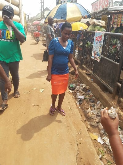 The image depicts an active street market scene in Nnobi, Nigeria, under daylight conditions.
In the mid-foreground, a woman, approximately 30-40 years old, is walking towards the viewer's left. She wears a blue, short-sleeved top with a white undergarment visible at the neckline, an orange knee-length skirt, and purple-striped flip-flops. Her hair is dark and short. She looks downwards and to her left.
To the woman's immediate right, an open drainage ditch or gutter runs parallel to the walking path, filled with accumulated refuse including plastic bags, wrappers, and various debris. The walking path itself is a light brown, packed dirt or concrete surface, showing a distinct shadow of a person in the foreground.
To the left of the primary subject, a man, approximately 30-50 years old, walks towards the right. He wears a green t-shirt with a colorful graphic print, dark trousers, and a black baseball cap with a white "NY" logo. He holds a white cloth or tissue to his face with his right hand. A partially visible leg and foot, wearing dark sandals, are present in the far left foreground.
Behind the woman and separating the pathway from the market stalls is a black metal fence. A white rectangular poster advertising "POS ISHALALALE" is attached to this fence. Beyond the fence, several market stalls are visible. One prominent stall is shaded by a blue and white striped umbrella, while another has a yellow umbrella. Piles of green leafy vegetables are displayed on a surface. A sign above one stall reads "POSPOS".
In the background, further down the street, more pedestrians, including a bald man in a grey t-shirt, and a motor scooter are visible amidst additional market structures. Utility poles and overhead wires are present against a bright, hazy sky. The overall environment indicates a bustling commercial area with visible waste accumulation along the drainage.