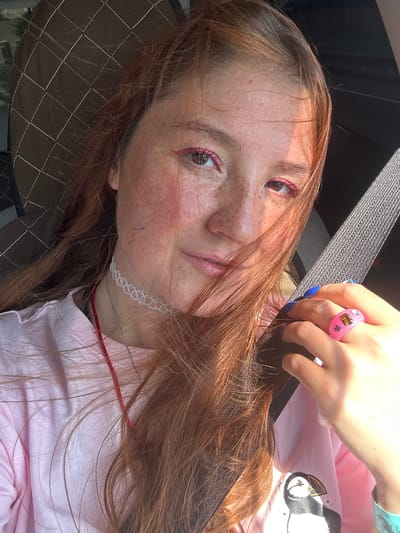 In this selfie, a young woman with red hair and freckles is seen from the chest up. She is wearing a pink t-shirt with a puffin graphic on it and a white choker necklace. Her makeup includes bright pink eyeliner. On her right hand, she wears a pink ring with gemstones and her nails are painted blue. The background shows a car interior, with a seatbelt and part of a headrest visible. The lighting suggests it is daytime, possibly with sunlight filtering through. The location is not specified as Крутобереговый, Russia, but the image is a close-up selfie taken within a vehicle.