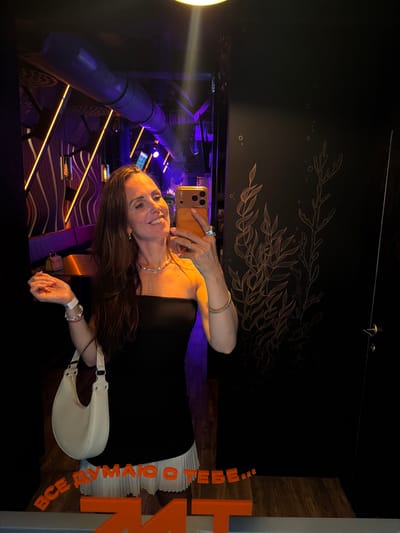 A woman is taking a selfie in a dimly lit interior space. She is wearing a black strapless dress and a pearl necklace, and is holding a smartphone in her right hand. She has long brown hair and is smiling at the camera. A light-colored shoulder bag is slung over her left arm.

The background is dark, with some illuminated elements visible. To the left, there is a large, curved, purple structure with metallic accents, possibly part of an installation or amusement ride. Yellow and purple lights are scattered throughout the scene, creating a vibrant and somewhat futuristic atmosphere. On the wall behind the woman, there is a delicate, line-art drawing of leaves and branches.

To the right, there is a wooden paneling and a large, orange, three-dimensional sign with Cyrillic letters that read "ВСЕГДА НАВСЕГДА". The overall impression is of an entertainment venue, perhaps an arcade, a theme park, or a nightclub. The lighting suggests it is nighttime or the indoor space is designed to be dark. The woman appears happy and confident.