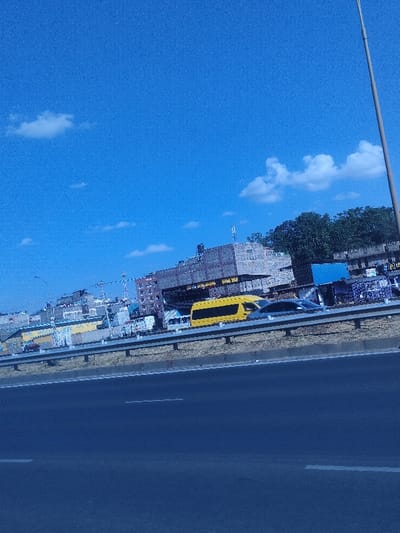 A daytime view presents an urban scene alongside a roadway. A yellow bus and a silver car are visible behind a metallic barrier. Several buildings of varying heights and constructions line the cityscape. The sky, predominantly blue, contains scattered clouds. A thin, tall pole is stationed on the right-hand side. The dark asphalt road occupies the lower portion of the image.