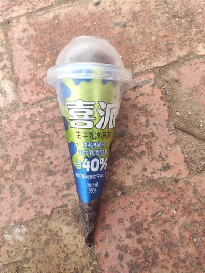 An ice cream cone with a clear plastic lid sits on a weathered red brick surface. The cone's wrapper is predominantly green and blue with white text. The text on the wrapper includes Chinese characters that translate to "Joy Pie," "Fresh Milk Ice Cream," and a percentage indicating "≥40% fresh milk content." There is also text that translates to "contains Oreo cookie crumbs" and "Net weight 95 grams." The bottom of the cone is dark brown and appears to be made of wafer. The background is a textured red brick walkway with some scattered dirt and a small twig. The image was taken in Jianyang City, China.