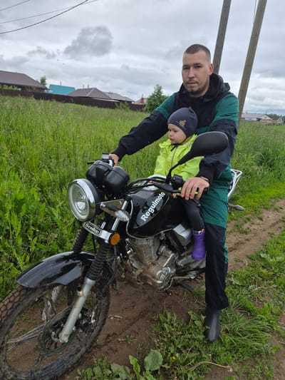 The image displays an adult male and a young child seated on a black "Regulmoto" motorcycle in a rural or semi-rural setting in Kharnavy, Russia. The motorcycle is positioned on a muddy dirt path, bordered by tall, unkempt green grass and scattered weeds.
The adult male, with short buzzed hair and light facial stubble, wears a black and dark green hooded jacket, matching dark green pants, and dark grey rubber boots. He is seated on the motorcycle, holding the handlebars with both hands, and looks directly at the viewer with a neutral expression. His left foot rests on the muddy ground.
A young child, likely a toddler, sits in front of the adult male on the motorcycle. The child is facing towards the left side of the frame, looking forward, and wears a neon yellow jacket, a dark grey beanie adorned with a light brown bear emblem, dark grey leggings, and bright purple patterned rubber boots.
The black motorcycle has the brand name "Regulmoto" visible on its fuel tank and "Senke" on the chrome bracket above the round headlight. The front wheel, fender, and lower engine components are splattered with mud. The motorcycle features knobby tires and a visible front disc brake.
The background shows an overcast sky with grey clouds, where three small, dark, distant airborne objects are faintly discernible. Two leaning wooden utility poles with overhead power lines stand behind the subjects. Further in the distance, a dark brown fence separates the field from several single-story houses with brown and blue roofs.