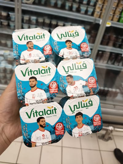 In a grocery store aisle, likely in Cebalat, Tunisia, a person's hand holds up a stack of six yogurt containers. The containers feature cartoon illustrations of young men in white and red sports jerseys. Each container has the brand name "Vitalait" printed in green and yellow, along with text in Arabic and French, indicating "Depuis 1998" (Since 1998). The illustrations depict three distinct individuals, each appearing twice on the stacked containers. The background shows rows of other yogurt products on shelves, partially blurred. Red and white confetti is depicted on the packaging. One of the containers also shows a circular logo with a Tunisian flag and the text "Sponsor Officiel" (Official Sponsor). The lighting suggests an indoor, well-lit environment typical of a supermarket.