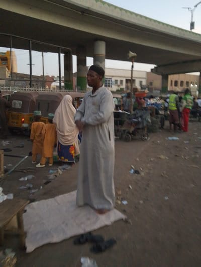 The image depicts an outdoor scene under a concrete overpass or bridge, likely in Sabon Gari West, Nigeria, during what appears to be late afternoon or early evening. The main subject is a man in the foreground, dressed in a light-colored, long traditional garment and a dark cap, standing barefoot on a white prayer mat. His hands are clasped, and he appears to be in a posture of prayer or contemplation.

Around him, the environment is bustling. To the left, a woman in a light-colored hijab and a patterned skirt stands with two young children, both dressed in matching orange-brown traditional outfits and small caps. Several brown three-wheeled vehicles, common as taxis, are visible in the mid-ground on the left, with one yellow one directly behind the woman and children.

The ground is unpaved, dusty, and strewn with various pieces of litter and debris. In the background, numerous people are scattered, some standing, some appearing to be vendors or shoppers. Two individuals on the right are notable for wearing bright safety vests, one yellow and one green. A wooden bench or table is partially visible on the far left. A tall pole with a loudspeaker-like object attached stands in the mid-ground to the right. Buildings of varying styles can be seen in the distance beyond the activity. The overall impression is that of a busy urban or market area. No clear, legible text is visible in the image.