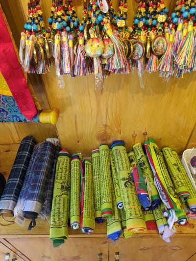 The image shows a close-up, eye-level, indoor shot of a display of Tibetan prayer flags and keychains. The prayer flags, mostly yellow with black Tibetan script and some imagery, are rolled up and arranged vertically in rows. They are tied with various colored rubber bands and some have colorful fabric borders. To the left of the prayer flags are two rolled-up items that appear to be umbrellas or fabric rolls with plaid patterns in shades of blue, black, and white. Above the prayer flags and umbrellas, a row of colorful beaded necklaces with small circular pendants and tassels hangs from a wooden surface. The background is a light-colored wooden wall with visible grain and knots. A small yellow object, possibly a toy or a decorative item, is attached to the wooden wall near the top left. The overall scene suggests a retail environment, likely a shop selling Tibetan handicrafts in Dharamshala, India, a city known for its Tibetan community and Buddhist culture. There are no people or animals visible in the image.