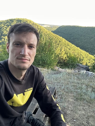 A man with short brown hair and stubble is sitting on a wooden bench in an outdoor setting. He is wearing a black long-sleeved shirt with a yellow graphic of a bat symbol and the text "THE ORIGINAL" on the left chest. The man is looking towards the camera. The background consists of rolling, tree-covered hills bathed in sunlight, with some dry grass and rocks in the foreground. The location appears to be a natural landscape, possibly on the outskirts of Pryvorottia Druhe, Ukraine.