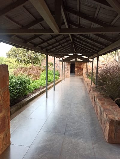 An outdoor corridor stretches into the distance, covered by a wooden roof structure. The corridor floor is composed of large grey tiles. One side features a raised brick edge with tall grass growing on top, while the other has a lower brick border with green foliage and trees visible beyond. Metal posts support the roof along the corridor's length. A dark opening can be seen at the corridor's end.