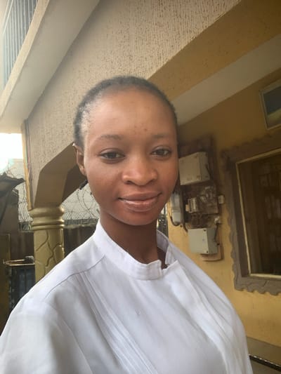 A portrait of a young Black woman, shown from the chest up, is captured in an outdoor setting. She is looking directly at the camera with a gentle smile. She is wearing a white collared shirt with decorative stitching. The background includes architectural elements, possibly a building exterior, with a rough-textured wall and some electrical fixtures visible. The lighting suggests it is daytime, with natural light illuminating the scene. The overall mood is relaxed and friendly. The image was taken in Shagamu, Nigeria.