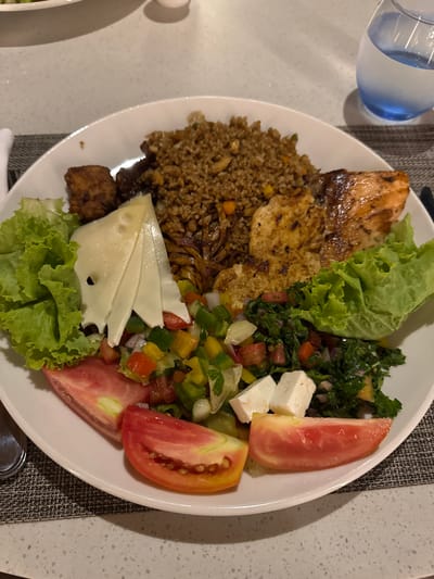 A plate of food features a diverse mix of ingredients. There's grilled chicken and fried rice, paired with what appears to be cheese slices and lettuce. A vibrant salad of diced tomatoes, bell peppers, and herbs sits beside slices of tomato and feta cheese. The plate offers a combination of protein, grains, and fresh vegetables. A glass of water is near the plate.