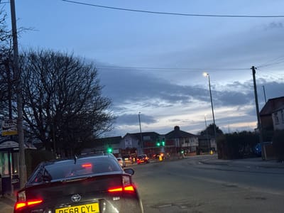 The image captures an urban street scene at dusk, specifically looking eastbound on Watling Street Road, Preston, United Kingdom, towards its junction with Victoria Road/St Gregory Road. The sky is overcast with a blend of dark blue and grey clouds. Ambient light is low, necessitating the illumination of artificial light sources.
In the immediate foreground, the rear of a dark-colored Toyota Prius (UK registration plate PF68 CYL) is visible, with its red brake lights illuminated and amber turn signals active. The vehicle is positioned in a traffic lane on a damp asphalt road surface, suggesting it is either stopped or decelerating. To the left of the foreground vehicle, a glass and metal bus stop shelter is partially visible, behind which stands a large, bare deciduous tree with extensive branching.
Further down the street, at a visible intersection, several vehicles are present, illuminated by their headlights and taillights. Multiple traffic signals at this junction display green lights, indicating active traffic flow. On the left side of the street, a building identified by green illuminated signage as "Marabana Balti" is visible. Beyond this, residential buildings, primarily terraced or semi-detached structures, line both sides of the roadway. Several tall, illuminated streetlights are positioned along the street, and utility poles with overhead wires are present. A dense green hedgerow borders the right side of the street. No discernible individuals are visible within the frame.