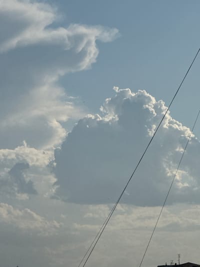 A low-angle, vertical shot captures a sky filled with clouds and two dark, thin lines that appear to be wires. The clouds are mostly white and puffy, with some areas of shadow indicating depth. The sky is a light blue. In the bottom right corner, a sliver of a dark roof with antennas is visible, suggesting an urban or suburban environment. The overall scene is serene and observational. The image is taken from the city of Abovyan, Armenia, looking upwards at the sky.