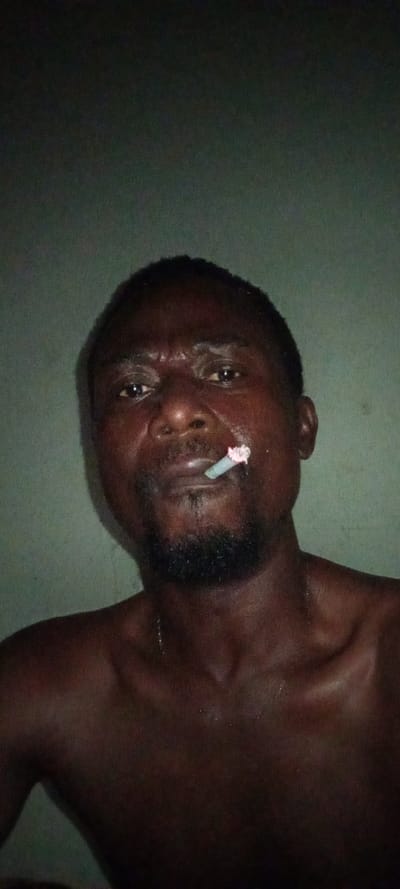An unadorned photograph presents a shirtless, dark-skinned man with a short beard and a cigarette held between his lips. His expression is intense, with furrowed brows and a direct gaze. The setting appears to be indoors, indicated by a plain, somewhat faded wall in the background. The image is cropped closely, emphasizing the man's face and upper torso. Sweat or moisture glistens on his skin.