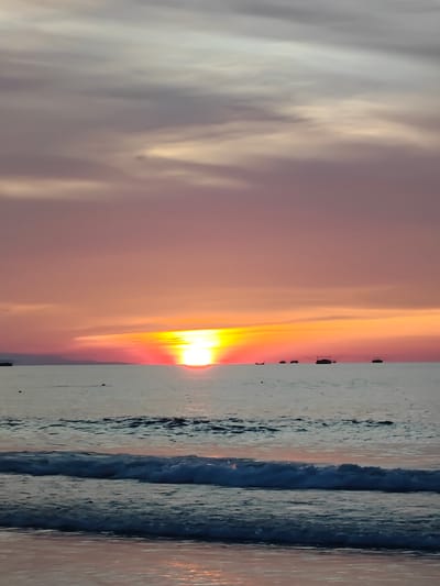 This image captures a serene sunset over the ocean at Curehtunong, Indonesia. The sky is a canvas of soft, diffused colors, transitioning from pale grey and dusty rose at the top to vibrant hues of orange and red as it meets the horizon. Wispy clouds add texture to the sky, catching the last rays of the sun.
The sun, a brilliant orb of yellow and red, is positioned just above the horizon, casting a warm glow across the water. The ocean itself is calm, with gentle waves rolling towards the shore. The foreground features a close-up view of these waves, their white foam contrasting with the darker, deeper blues and grays of the water. The wet sand at the very bottom of the frame reflects the sunset's colors, adding a subtle pinkish hue.
In the distance, several small boats are visible on the water, silhouetted against the vibrant sky. These boats suggest human activity and perhaps local fishing or transportation. The overall atmosphere is one of tranquility and natural beauty, evoking a sense of peace and the end of a day. The time of day is clearly sunset, indicated by the sun's position and the warm, rich colors in the sky and on the water. There is no visible text in the image.