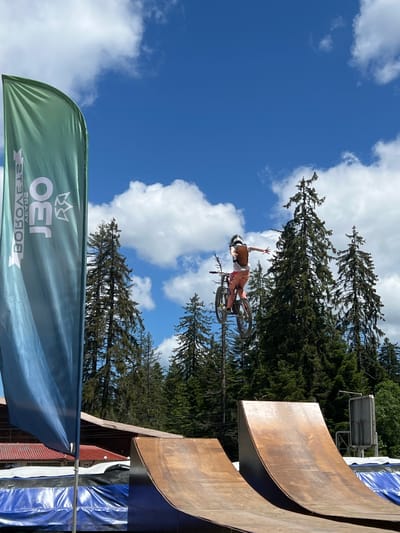 A person on a mountain bike is captured mid-air during a jump. The rider, wearing a helmet and a backpack, is dressed in orange pants and a light-colored shirt. They are positioned against a backdrop of tall, dark green evergreen trees and a bright blue sky with scattered white clouds. In the foreground, two large, curved ramps made of wood are visible. To the left, a tall, green flag with white text and a logo stands prominently. The scene appears to be an outdoor sports venue, likely a bike park or a competition site. The specific location in Samokov, Bulgaria, is not identifiable from the visual information alone, but the natural setting with evergreen trees suggests a mountainous or hilly area.