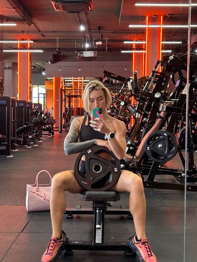 A woman is sitting on a weight bench in a gym, taking a selfie with her phone. She is holding a weight plate with the number "12" on it in her lap. She is wearing a black sports bra, black shorts, and pink and black athletic shoes. A pink gym bag is placed on the floor next to the bench. The gym has orange neon lighting and numerous weight machines visible in the background. The woman has blonde hair and visible tattoos on her arms. She is wearing a watch on her left wrist. The location is identified as a gym in Chalong, Thailand.