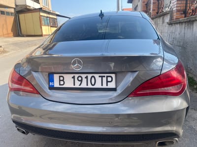 The image displays the rear of a grey Mercedes-Benz CLA-class sedan, parked on a light-colored paved surface in Vetren, Bulgaria. The vehicle's Bulgarian license plate, "B 1910 TP," is centrally mounted on the trunk lid, below the prominent Mercedes-Benz emblem. The rear window is tinted and appears dusty. Red taillight assemblies flank the trunk lid. The lower rear bumper incorporates a black mesh diffuser with dual chrome exhaust tips and two circular parking sensors. To the left of the vehicle, an upward-sloping road leads past a two-story building featuring a brown ground-floor garage door and a lighter-colored upper level, adjacent to a smaller, yellow-paneled ancillary structure with multiple windows. To the right, a low brick wall topped with black decorative wrought iron fencing separates the vehicle from an adjacent property, where a portion of a building with a red tile roof and minimal greenery is visible. The scene is illuminated by direct sunlight under a clear blue sky. No individuals or active events are depicted.