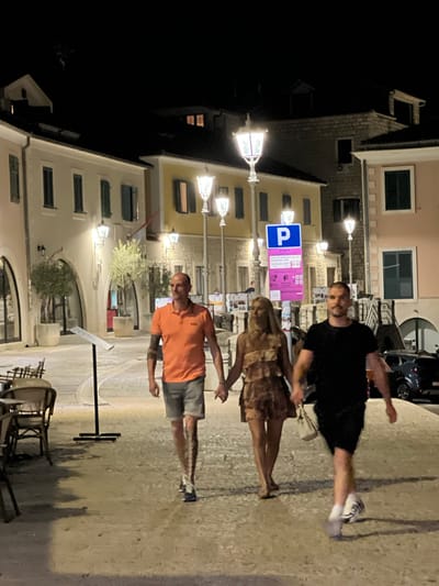 Three people are walking on a cobblestone street at night in Herceg Novi, Montenegro. The person on the left is a bald man wearing an orange polo shirt and grey shorts. He has tattoos on his left arm and is walking towards the viewer. In the center, a woman with blonde hair is wearing a light brown, tiered dress and is holding hands with the man on the right. She is also walking towards the viewer. The man on the right has short hair and a beard and is wearing a black t-shirt and black shorts. He is carrying a white purse in his right hand and is walking towards the viewer. Behind the group, there are buildings with lights on. A blue parking sign is visible on the right side of the image. The ground is uneven and appears to be sand and cobblestone. There are cafe tables and chairs on the left side of the image.