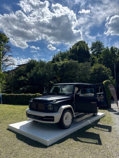 A dark-colored SUV, with white wheel arches and wheels, is parked on a white platform. The platform sits on a patch of grass. The car's doors are open. In the background are trees and a building. Above, the sky is blue with fluffy white clouds. A banner stands next to the car. The location is Como, Italy.