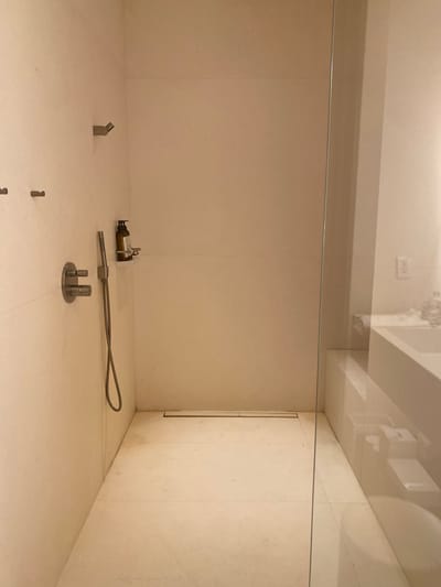 This image appears to be a photograph taken inside a minimalist, possibly institutional or museum-like space. The scene is dominated by light-colored walls with subtle vertical seams, suggesting large panels or tiles. A horizontal metal rod spans the upper portion of the frame, intersecting where the side walls meet the back. A glass ceiling or barrier is visible in the upper portion of the image.
On the floor, several small, metal objects are arranged. From left to right: a small angled piece, a collection of what appears to be an amber colored liquid dispensing bottle next to a complex piece of machinery, then a flexible metal tube attached to a cylindrical component, and two small cylindrical pieces protruding from a circular base. The walls are off-white with subtle seams, suggesting a simple background.
There are no people visible in the frame. There are no actions or interactions to describe based on the objects present.
There is no indication of a location in the city of El Caracol, Mexico, unless these objects are known to be an art installation or artifact exhibited in that specific locale.