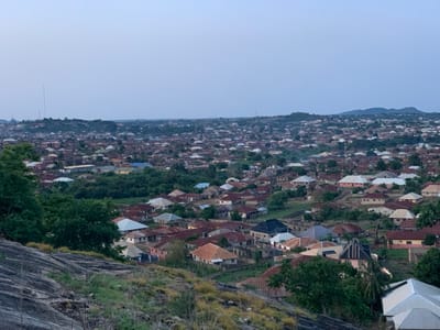 The image depicts a dense urban landscape in Kabba, Nigeria, viewed from an elevated position. The scene is dominated by closely packed buildings, predominantly single-story residential structures with diverse roof colors and materials including terracotta and metal sheeting. Interspersed among the houses are patches of green vegetation, with trees and shrubbery evident in both the foreground and scattered throughout the city. A rocky, grass-covered terrain forms the immediate foreground, suggesting the vantage point is from a hillside or outcrop. In the distance, the city extends towards the horizon, where a line of hills is visible. The sky is a pale blue, indicating either early morning or late afternoon light. Tall communications towers are visible in the distance. The overall impression is of a sprawling, densely populated urban environment set within a natural, hilly landscape.
