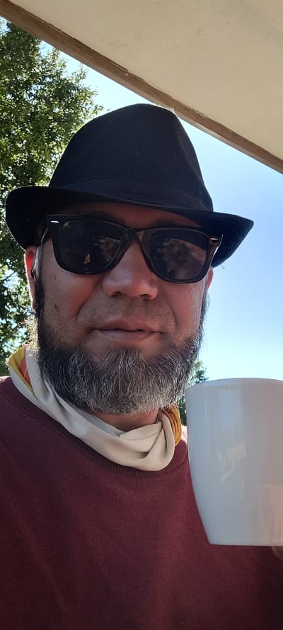 In Milisi, Georgia, a man is pictured close-up, holding a white coffee mug. He is wearing a black fedora, sunglasses, a maroon sweatshirt, and a light-colored scarf around his neck. His beard is gray and neatly trimmed. The background shows trees and a clear blue sky, suggesting an outdoor setting. The man appears to be relaxed, possibly enjoying a beverage outdoors.