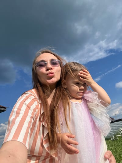 A woman wearing sunglasses and a peach and white striped shirt is taking a selfie outdoors. She is puckering her lips. A young girl in a pastel colored dress is standing next to her, with her hand on her forehead. The background is a blue sky with some clouds. The woman is in the foreground, and the girl is to her right. The scene appears to be in the city of Kharnavy, Russia.