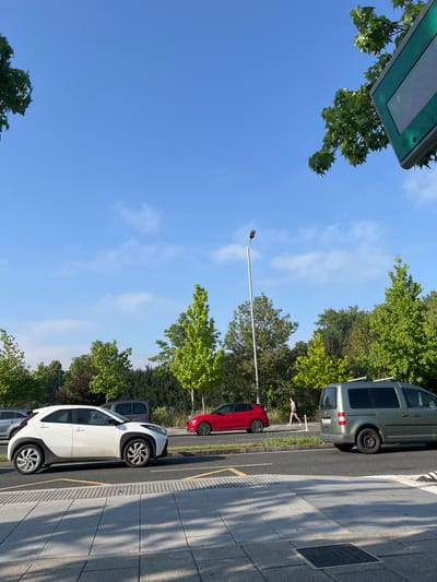 The image depicts a street scene in Vitoria-Gasteiz, Spain, under a clear blue sky with scattered white clouds. In the foreground, a light-colored, modern hatchback is parked on a street with yellow line markings. Adjacent to it, a silver minivan is also present. A person in casual attire is walking along the side of the road, moving from right to left within the frame.

The street is bordered by a sidewalk made of grey paving stones, with shadows cast by unseen objects. Beyond the street, a line of trees with vibrant green foliage forms a backdrop, with a tall lamppost positioned between the trees and the road. In the distance, more trees and some structures are visible. The overall scene suggests a typical urban environment during daylight hours.