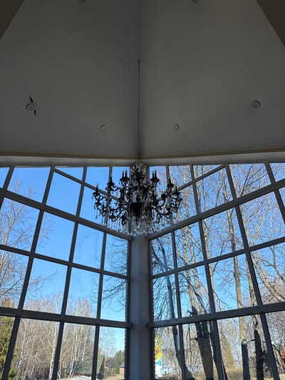 The image shows an interior view with a large chandelier hanging below a grid-patterned glass ceiling. Outside the glass panels, trees are visible against a clear blue sky. The surrounding architecture includes a smooth, white ceiling with recessed lighting. The scene appears to be an atrium or sunroom-like space. There is no evidence to suggest that it is in Galevo, Russia. The image does not include people or specific actions.