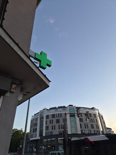 This low-angle, upward-looking photographic image captures a late afternoon or early evening urban scene in Esch-sur-Alzette, Luxembourg, focusing on architectural elements and the sky.
In the left foreground, a portion of a dark gray, textured building facade with a prominent overhang is visible. Attached to the underside of this overhang, extending outwards, is a large, green, three-dimensional cross sign, illuminated by numerous small, individual LED lights, characteristic of a pharmacy. Below the overhang, on the building's exterior, an electrical box assembly is mounted, featuring a red-and-white labeled component reading "NEON alarm system" above a white button or switch. A metallic gutter runs along the top edge of the visible building section.
The background is dominated by a distinctive, modern, multi-story building with a rounded or semi-circular primary footprint. This structure features a facade composed of alternating sections of light gray and darker gray panels, interspersed with numerous rectangular windows, many equipped with external blinds or shutters. A vertical strip of transparent glass extends upwards on the building's curved face. The upper levels of this building are set back, and some glass surfaces reflect the warm, yellowish light of the setting sun on the right side.
Below the main curved structure, at ground level, are commercial storefronts with visible signage. Signs include "ACM" repeated multiple times, "EXPO GOMA," "PORTES ET FENETRES," "ENTREE," and what appears to be "BETALICA." A street pole supporting multiple traffic lights stands in front of this building. A dark-colored vehicle, likely a bus or tram, is partially visible in the lower right, obstructing the view of the building's base; a small red illuminated pedestrian signal is visible on its side. To the left of the curved building, a cluster of green deciduous trees is visible.
The upper portion of the image is filled with a clear, light blue sky, featuring a few faint, wispy clouds, predominantly in the upper left quadrant. Towards the right horizon, the sky transitions to a warmer, yellowish-orange hue, indicating either sunrise or sunset. A faint, linear contrail from an aircraft is discernible in the far upper right sky. No individuals are clearly visible or identifiable in the scene.