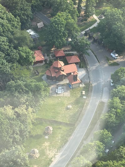 This aerial shot shows the vicinity of the Monastery of the Holy Archangels Gabriel in the city of Vozdovac Urban Municipality, Serbia. A building complex with multiple structures, distinguished by their red tile roofs, sits among lush greenery. Adjacent to the building complex is a paved road that curves along the side. A few vehicles can be seen on a connecting road to the main street, and a statue rests on the grass. There are also two gazebos. Surrounding the Monastery are trees, which are located on all sides of the complex. The image suggests a serene environment situated amidst natural landscape.