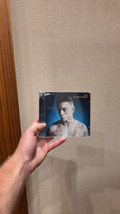 A hand holds a CD case in front of a textured, beige wall, with a wooden surface visible at the bottom of the frame. The CD cover features a stylized image of a person's upper body, appearing to be a statue with marble-like skin and cracks filled with gold. The person's head is bowed, and their eyes are closed, conveying a sense of repose or introspection. The background of the CD cover is a dark, gradient blue, suggesting a night sky or deep water. Text on the left side of the cover reads "I.N.L." and "KLASSA MODE" in a gold, vertical font. The lighting in the image is moderate, casting soft shadows, and it appears to be indoors during the day. The setting of Bania, Bulgaria, is not explicitly depicted in the image itself.