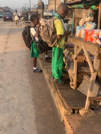 The image captures a street scene in Shagamu, Nigeria, with two young boys standing by a roadside stall. The boy on the left, wearing a white t-shirt and green skirt, has a large black backpack. The boy on the right, in a yellow shirt and green trousers, is also carrying a backpack and appears to be looking at items on the stall, which displays various packaged goods, including bread. A man in a white shirt and brown trousers is walking away from the stall down the dusty road. In the background, a dark-colored van is parked on the side of the road, and other people and buildings are visible, suggesting a bustling urban environment. The time of day appears to be late afternoon, as indicated by the warm, golden light casting long shadows.