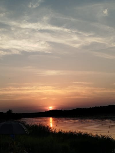 A sunset over a body of water is depicted. The sky is filled with clouds in shades of blue, gray, and orange. The sun is a bright orange orb low on the horizon, casting a shimmering reflection on the rippling water. A silhouetted treeline forms the background of the scene. In the foreground, tall grasses and a dark-colored umbrella suggest a lakeside or riverside setting. There are no people visible in the image. The location is specified as Topolina, Poland.