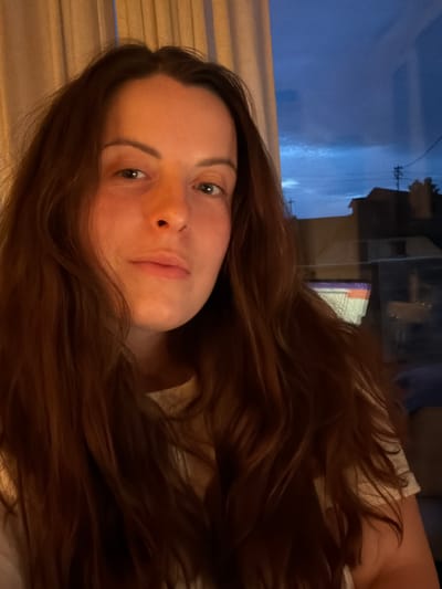 A young woman with light skin and long, auburn-colored hair is seen in an indoor setting. She is facing the camera with a neutral expression. Outside the window behind her, there is an urban scene, possibly in Gangloff, Germany, with a glimpse of buildings and overhead electrical wires. The lighting suggests it is either dusk or dawn. The image is rotated, with the horizontal orientation of the subject appearing vertical.