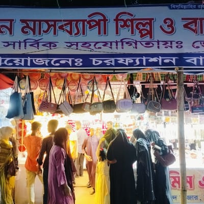 The image captures a bustling night market scene at a brightly lit stall in Charfasson, Bangladesh. Numerous people, including men, women (many wearing hijabs or abayas), and a child, are gathered, seemingly browsing or shopping. Their faces are largely obscured or turned away from the camera, adding to the candid feel of the moment.

The main display of the stall features a wide array of handbags and purses of various styles, colors, and sizes, hanging from racks. Some bags bear text like "HELLOT". In the background, beyond the handbags, other items, possibly hats or decorative goods, are visible. The bright artificial lighting of the stall creates a warm glow and some glare, contrasting with the presumed darkness of the night outside.

A large banner hangs above the stall with Bengali text. The readable parts include "বিস্মিল্লাহির রাহ" (Bismillahir Rahman), an Islamic phrase meaning "In the name of Allah, the Most Gracious, the Most Merciful." Other sections indicate "ন মাসব্যাপী শিল্প ও বা" (likely "Month-long industry and trade fair") and "আয়োজনেঃ চরফ্যাশন বা" (organized by Charfasson, likely "Charfasson Market" or "Charfasson Commercial"). This suggests the stall is part of a larger event, possibly a month-long trade fair or market organized in Charfasson. The overall impression is one of active commerce and community gathering in a vibrant market setting.