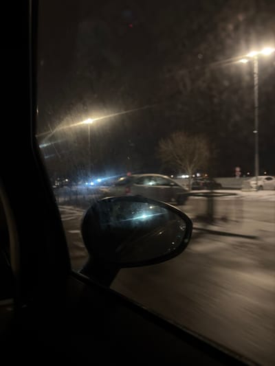A nighttime scene from inside a car. A side mirror reflects a bright light source, likely another vehicle. Rain or moisture obscures the windshield, causing light refraction. Outside, streetlights illuminate parked cars on the side of the road and a bare tree. The overall ambiance is dark and wet, captured in a slightly blurred perspective.