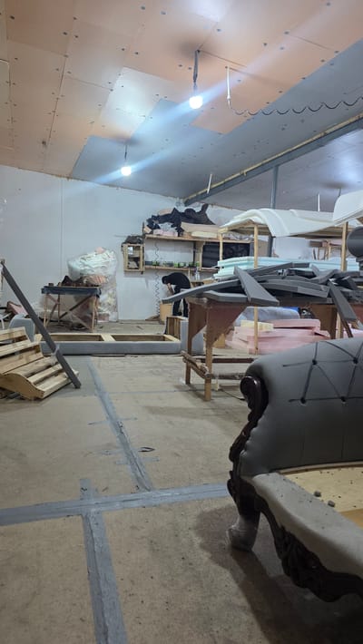 This image depicts an indoor workshop, likely a furniture upholstery or manufacturing space, with materials and partially finished items scattered around. The setting appears somewhat unorganized, with various pieces of foam padding, fabric scraps, and wooden frames visible. In the background, a person wearing a dark shirt and pants is bent over a workbench, seemingly engaged in work. To the right, the leg and base of an upholstered furniture piece with decorative carving are prominent. The walls are covered with large panels, some appearing to be insulation, and industrial-style lighting fixtures illuminate the space. The floor is a concrete or cement surface, with some areas taped together. The overall impression is of an active workspace where furniture is being constructed or repaired. There are no discernible clues about the time of day or weather conditions. No visible text is present in the image.