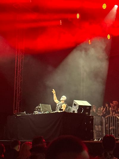 The image captures a DJ performing on a stage bathed in red light, with smoke creating a dramatic atmosphere. The DJ, wearing a white mask and a dark outfit, is seen raising their right hand. In front of the DJ is a table with audio equipment, including speakers and a laptop. A crowd is visible behind a metal barrier in the background, some holding up their hands. The stage is supported by metal scaffolding. The scene appears to be an indoor concert or event, possibly within Palilula Urban Municipality, Serbia, given the context provided.