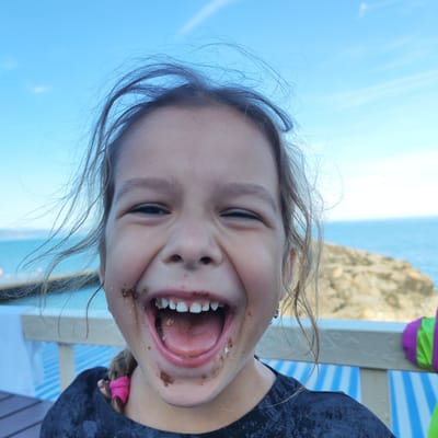 A close-up, eye-level medium shot captures a young girl with dark hair, windblown and messy, laughing exuberantly outdoors. Her mouth is wide open, revealing her teeth and tongue, and she has traces of dark food, likely chocolate, smeared around her mouth and chin, suggesting she has recently eaten something messy. Her eyes are squinted with joy, and the overall expression is one of uninhibited happiness.
The background indicates a bright, sunny day with a clear blue sky. Beyond the girl, the turquoise expanse of the sea stretches to the horizon, meeting a light-colored coastline with rocky formations. A beige-colored railing or barrier is visible in the foreground, along with striped blue and white surfaces, possibly lounge chairs or a beach umbrella. The context of Alushta, Ukraine, suggests a seaside location, likely a beach or a promenade.